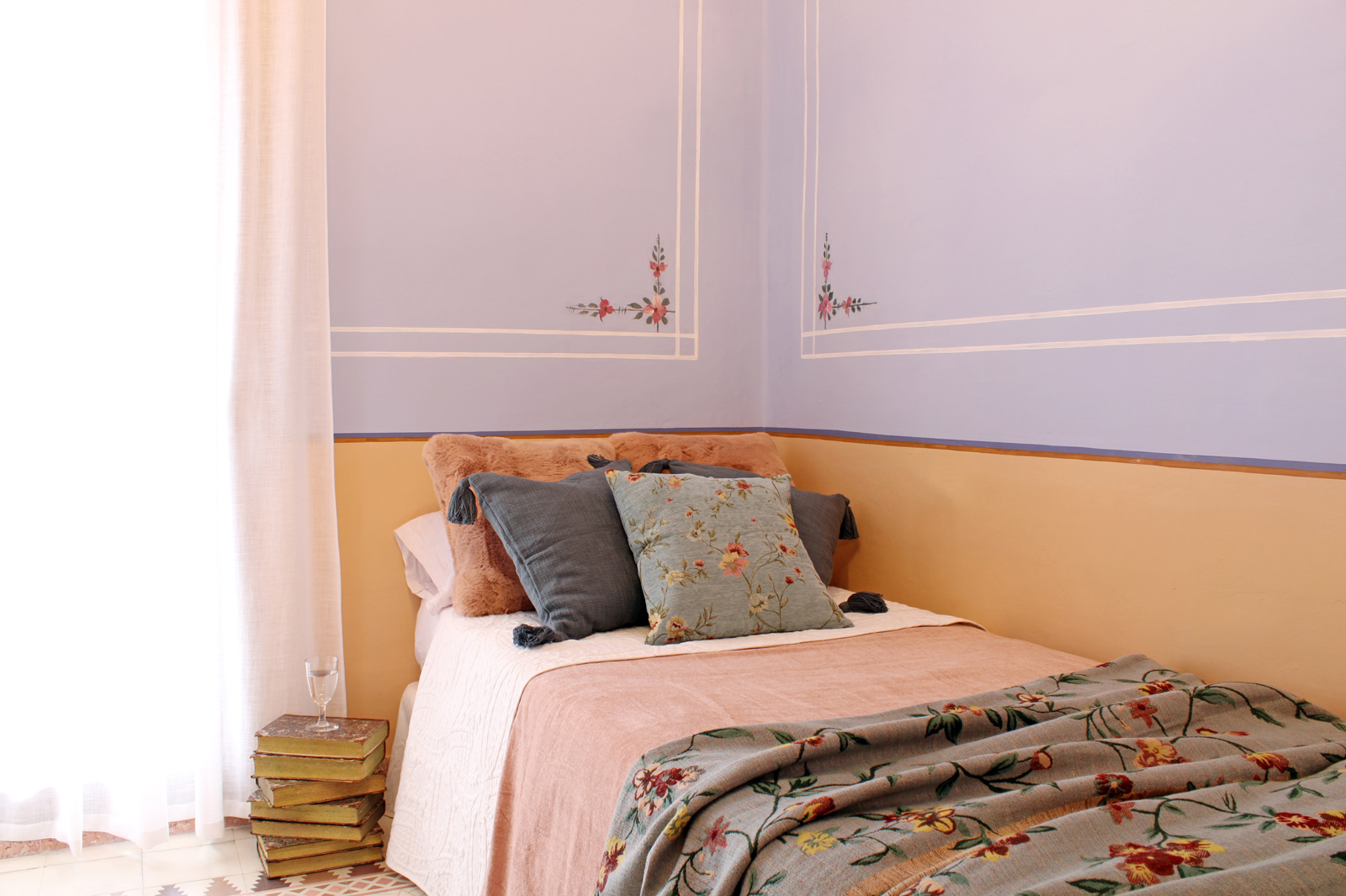 Detalle de dormitorio, esquina con cama con cojines en tonos rosas azules y floridos, montón de libros a modo de mesilla con copa encima